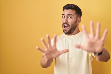 Handsome hispanic man standing over yellow background afraid and terrified with fear expression stop gesture with hands, shouting in shock. panic concept.