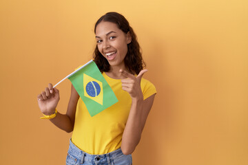 Young hispanic woman holding brazil flag pointing fingers to camera with happy and funny face. good energy and vibes.