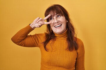 Middle age hispanic woman standing over yellow background doing peace symbol with fingers over face, smiling cheerful showing victory