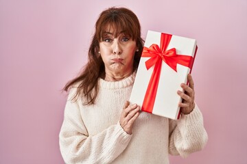 Middle age hispanic woman holding present puffing cheeks with funny face. mouth inflated with air, catching air. - obrazy, fototapety, plakaty