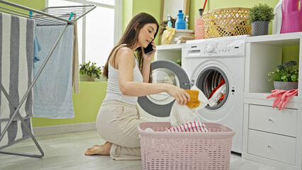 Young beautiful hispanic woman washing clothes talking on smartphone at laundry room