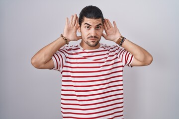 Fototapeta premium Hispanic man with long hair standing over isolated background trying to hear both hands on ear gesture, curious for gossip. hearing problem, deaf