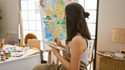 Beautiful, focused, young hispanic woman artist sitting on a chair, immersed in creativity, drawing in an art studio, indoors