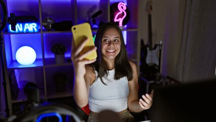 Fototapeta na wymiar Smiling young hispanic woman, streaming a thrilling video game, engrossed in digital entertainment from her neon-lit gaming room during a lively video call