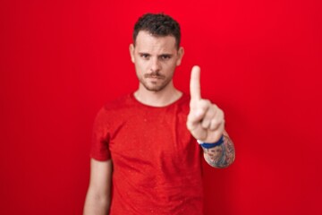 Young hispanic man standing over red background pointing with finger up and angry expression, showing no gesture