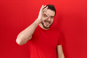 Young hispanic man wearing casual red t shirt doing ok gesture with hand smiling, eye looking through fingers with happy face.