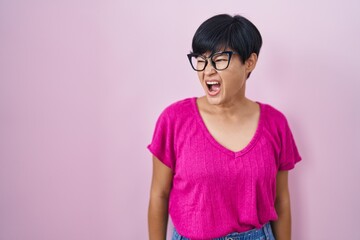 Young asian woman with short hair standing over pink background angry and mad screaming frustrated and furious, shouting with anger. rage and aggressive concept.