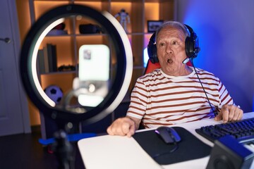 Senior man with grey hair recording gamer tutorial with smartphone at home scared and amazed with...