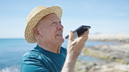 Senior grey-haired man tourist wearing summer hat sending voice message by smartphone at seaside