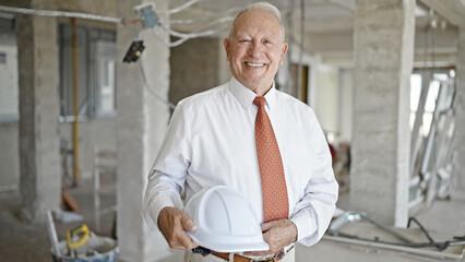 Senior grey-haired man architect smiling confident holding hardhat at construction site
