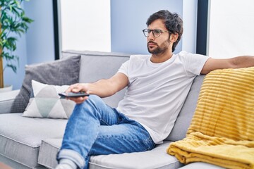 Handsome latin man holding television remote control skeptic and nervous, frowning upset because of...