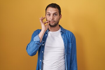 Hispanic man standing over yellow background mouth and lips shut as zip with fingers. secret and...