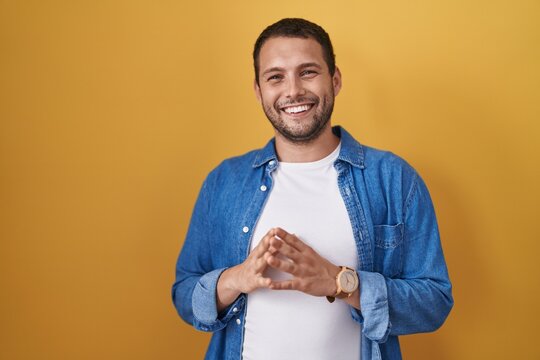 Hispanic man standing over yellow background hands together and fingers crossed smiling relaxed and cheerful. success and optimistic