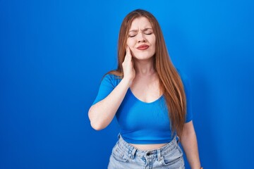 Redhead woman standing over blue background touching mouth with hand with painful expression because of toothache or dental illness on teeth. dentist