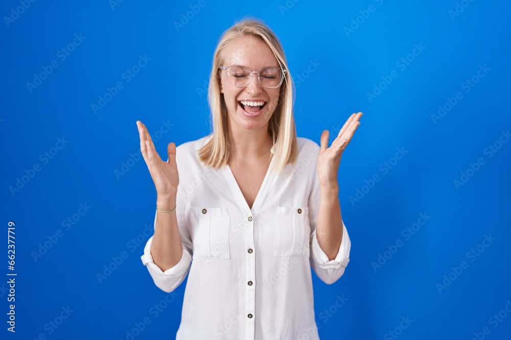 Sticker Young caucasian woman standing over blue background celebrating mad and crazy for success with arms raised and closed eyes screaming excited. winner concept