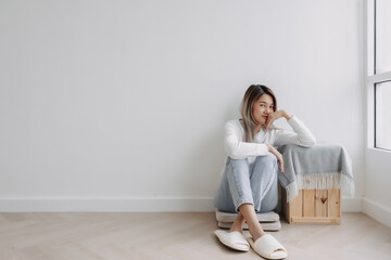Asian Thai woman sitting on floor next to the window, thinking of good thing memories and value time, resting chin on hand and put on table, chilling at apartment room in winter day alone.