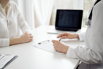 Fototapeta premium Doctor and patient sitting near each other at the white desk in clinic. Female physician is listening filling up a records form. Medicine concept