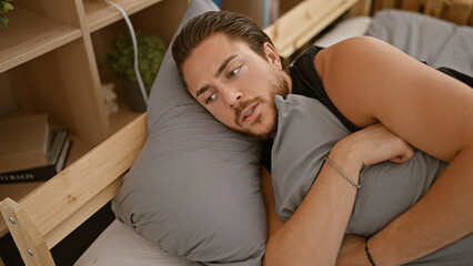 Fototapeta premium Young hispanic man lying on bed with sad expression hugging pillow at bedroom