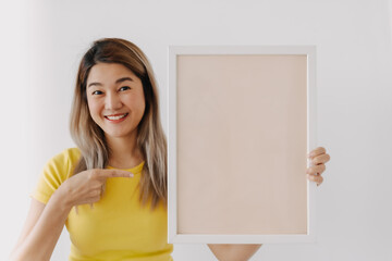 Thai asian woman wear yellow t-shirt, showing and pointing finger at empty white screen frame ,...