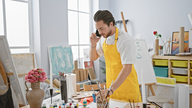 Young hispanic man artist talking on smartphone standing at art studio