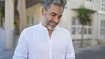 Young grey-haired hispanic man, confidently smiling, standing, basking in sunny street ambiance, a...