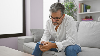 Fototapeta premium Young hispanic grey-haired man using smartphone sitting on sofa at home