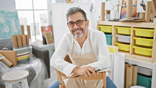 Grey-haired young hispanic man joyfully painting, a bearded artist with glasses, smiling confidently, sitting in an art studio holding a paintbrush