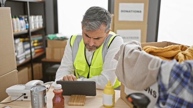 Attractive young hispanic man with grey hair, volunteering at a charity center, passionately works online via touchpad while sitting indoors amid a bustling warehouse.