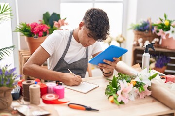 Young hispanic teenager florist using touchpad writing on notebook at flower shop