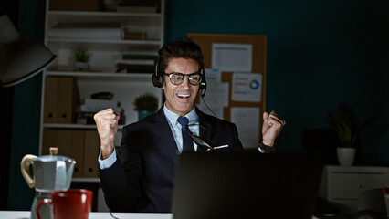 Young hispanic man business worker using laptop and headphones celebrating at the office