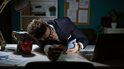 Young hispanic man business man tired leaning on table using smartphone at the office