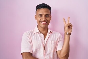 Young hispanic man standing over pink background smiling with happy face winking at the camera...