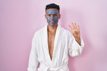 Young hispanic man wearing beauty face mask and bath robe showing and pointing up with fingers...