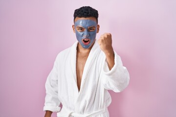 Young hispanic man wearing beauty face mask and bath robe angry and mad raising fist frustrated and...