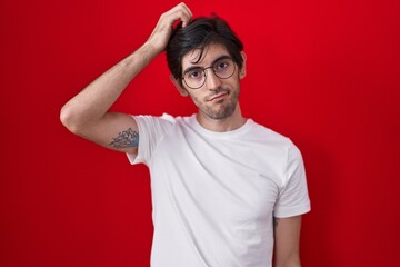 Young hispanic man standing over red background confuse and wondering about question. uncertain...