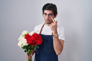 Young hispanic man holding bouquet of white and red roses pointing to the eye watching you gesture, suspicious expression