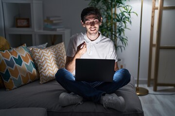 Young hispanic man using laptop at home at night doing money gesture with hands, asking for salary...