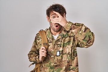 Hispanic young man wearing camouflage army uniform peeking in shock covering face and eyes with hand, looking through fingers with embarrassed expression.