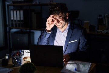 Hispanic young man working at the office at night doing ok gesture with hand smiling, eye looking through fingers with happy face.