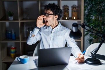 Hispanic man working at the office at night shouting and screaming loud to side with hand on mouth. communication concept.