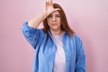 Young hispanic woman with red hair standing over pink background making fun of people with fingers...