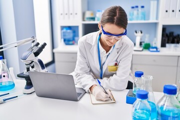 Young caucasian woman scientist using laptop writing on notebook at laboratory - obrazy, fototapety, plakaty