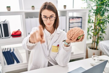 Young caucasian doctor woman holding brain as mental health concept pointing with finger to the camera and to you, confident gesture looking serious