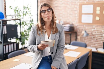 Young hispanic woman working at the office wearing glasses making fish face with lips, crazy and...
