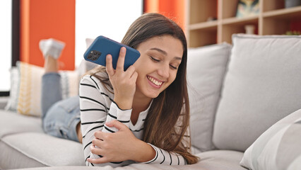 Young beautiful hispanic woman listening to voice message by smartphone lying on sofa at home