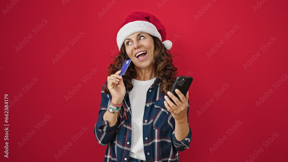 Poster Middle age hispanic woman shopping with smartphone and credit card wearing christmas hat over isolated red background