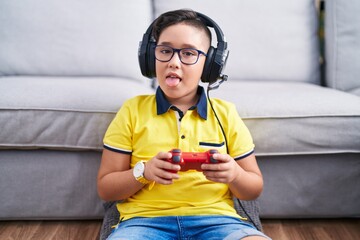 Young hispanic kid playing video game holding controller wearing headphones sticking tongue out happy with funny expression. emotion concept.