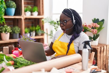Young african woman working at florist shop doing video call scared and amazed with open mouth for surprise, disbelief face - obrazy, fototapety, plakaty