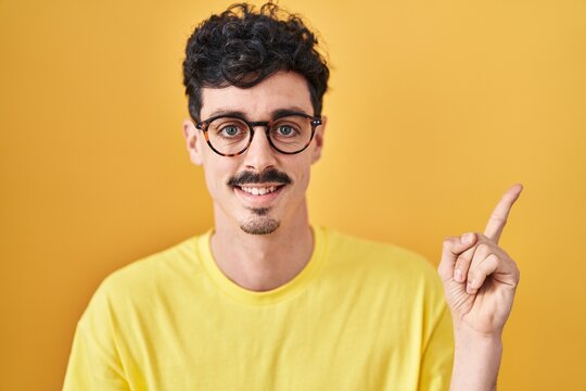 Hispanic man wearing glasses standing over yellow background with a big smile on face, pointing with hand and finger to the side looking at the camera.