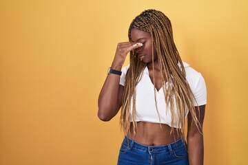 African american woman with braided hair standing over yellow background tired rubbing nose and eyes feeling fatigue and headache. stress and frustration concept.